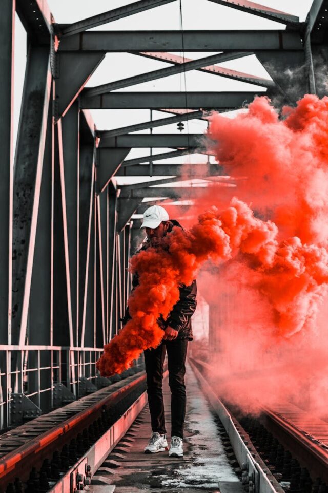 Man holding orange smoke bomb on railway bridge, creating a vibrant and dramatic scene.
