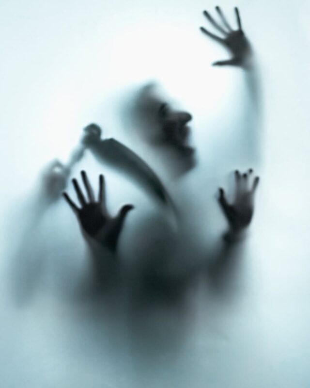 A mysterious silhouette with raised hands and a knife creates an eerie visual against frosted glass.