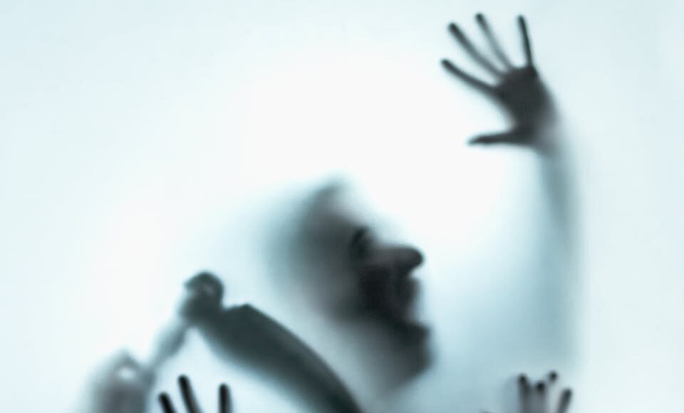 A mysterious silhouette with raised hands and a knife creates an eerie visual against frosted glass.