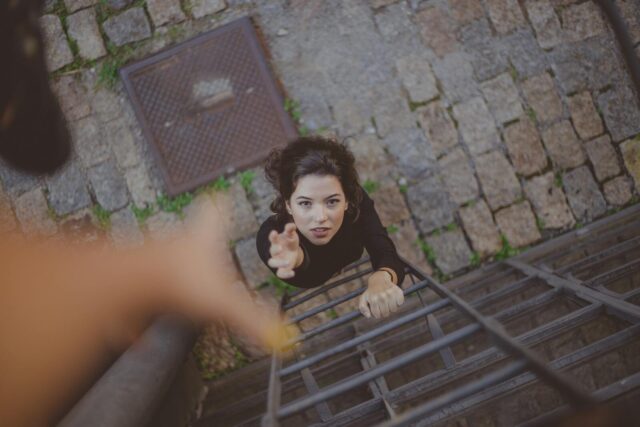 Photo by Samantha Garrote Woman climbs iron ladder outdoors, reaching for help with determination.