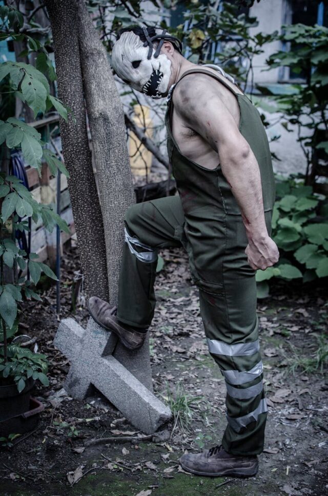 Photo by Gustavo Martínez Creepy masked figure standing next to a Christian cross in a garden setting.