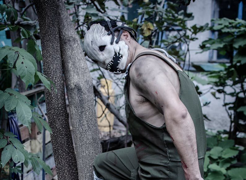 Creepy masked figure standing next to a Christian cross in a garden setting.