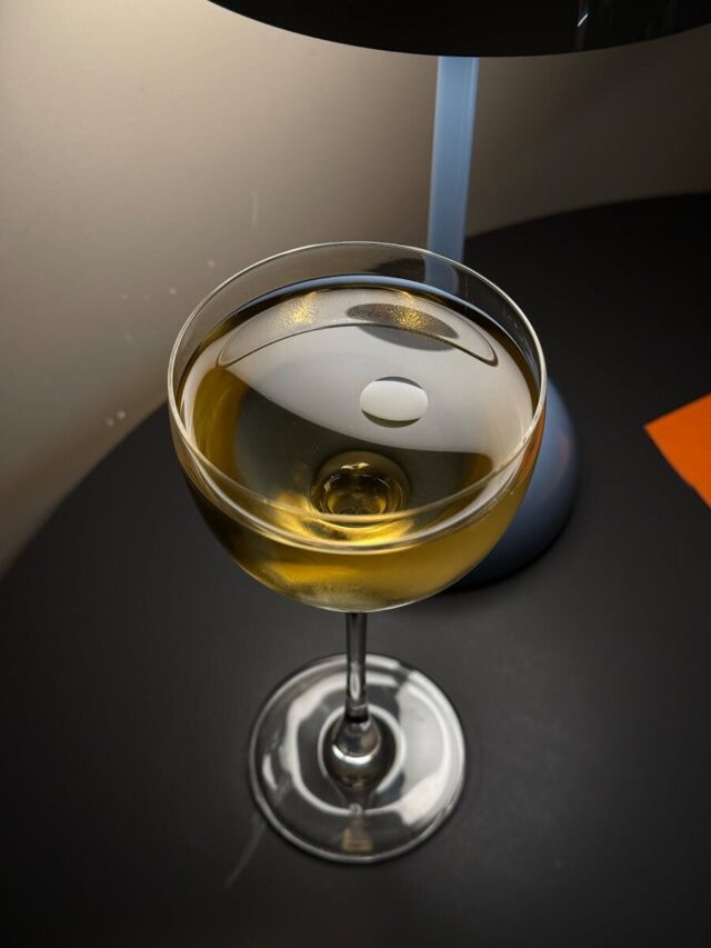 Photo by Yuliia Mrnk A glass of golden-colored liquid sits elegantly on a black surface, illuminated by soft warm light.