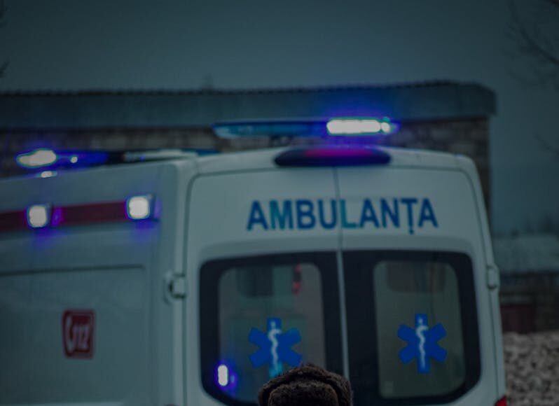 Photo by Gabriel Colta Elderly man walking in front of ambulance on city street during the evening.