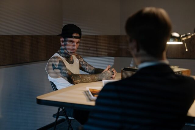 Photo by cottonbro studio Tattooed man in white tank top and cap during intense police interrogation in dimly lit room.