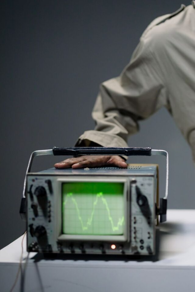 A hand adjusting an oscilloscope displaying a waveform, captured in a studio setup.