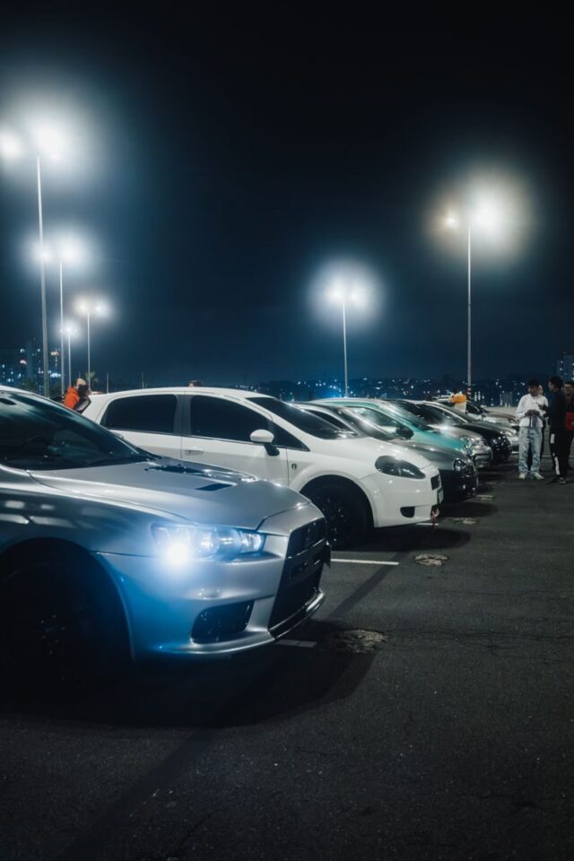 Photo by Matheus Bertelli A vivid night scene capturing a row of cars parked under bright streetlights in a spacious parking lot.