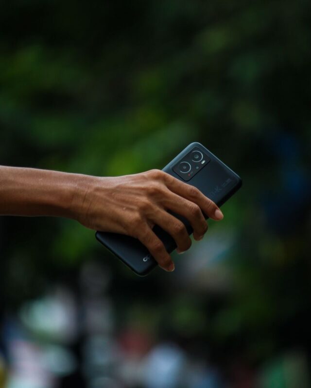 Photo by Vivek Solanki A person's hand holding a smartphone with a blurred outdoor background, focused view.