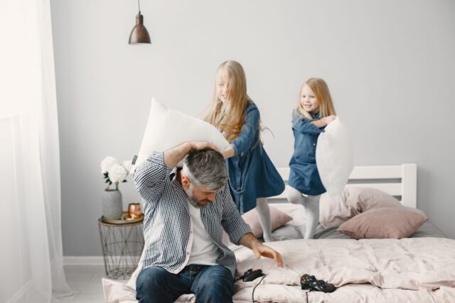 Photo by Gustavo Fring Father and daughters enjoy a playful pillow fight in a cozy bedroom setting.